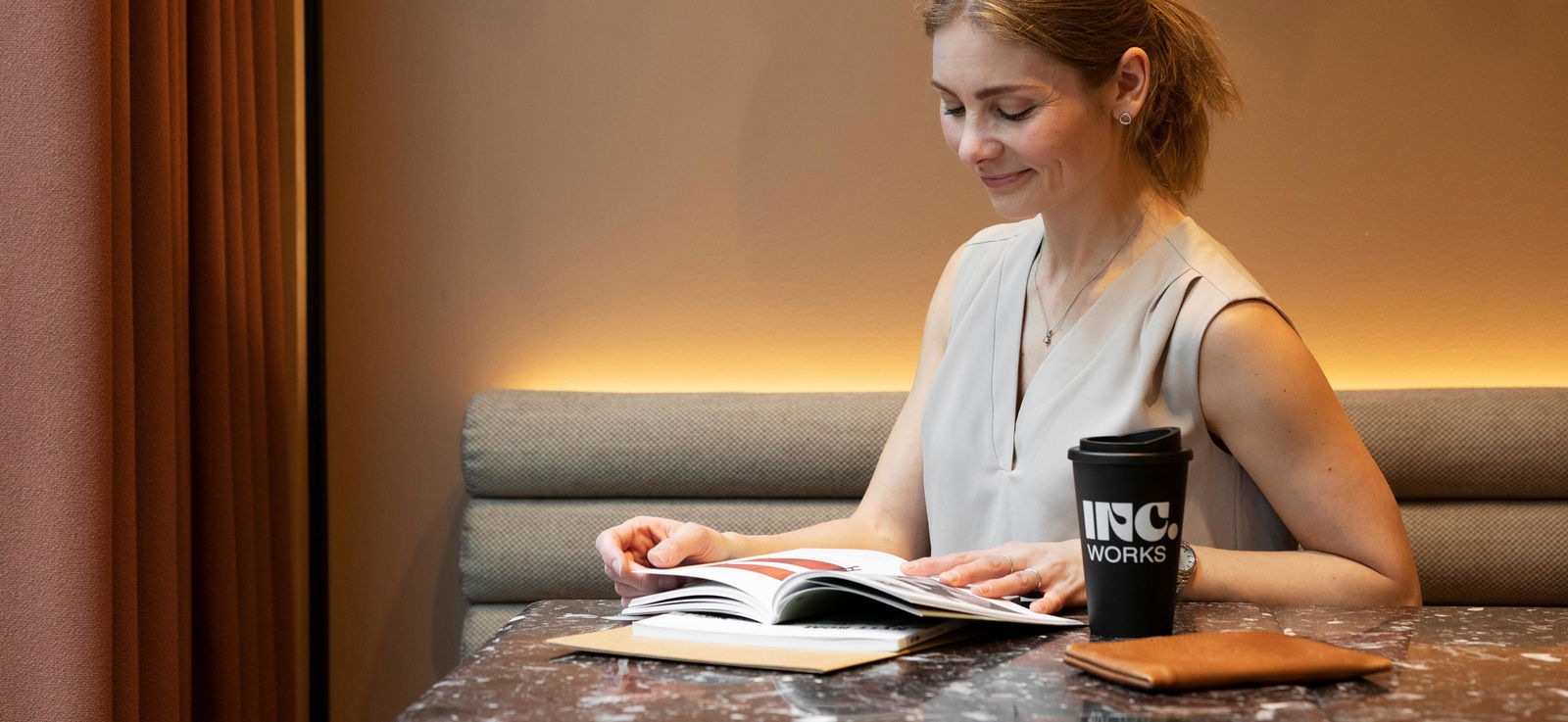 Kvinne sitter ved et bord og leser, og har kaffekopp ved siden av seg med INC.-logo på